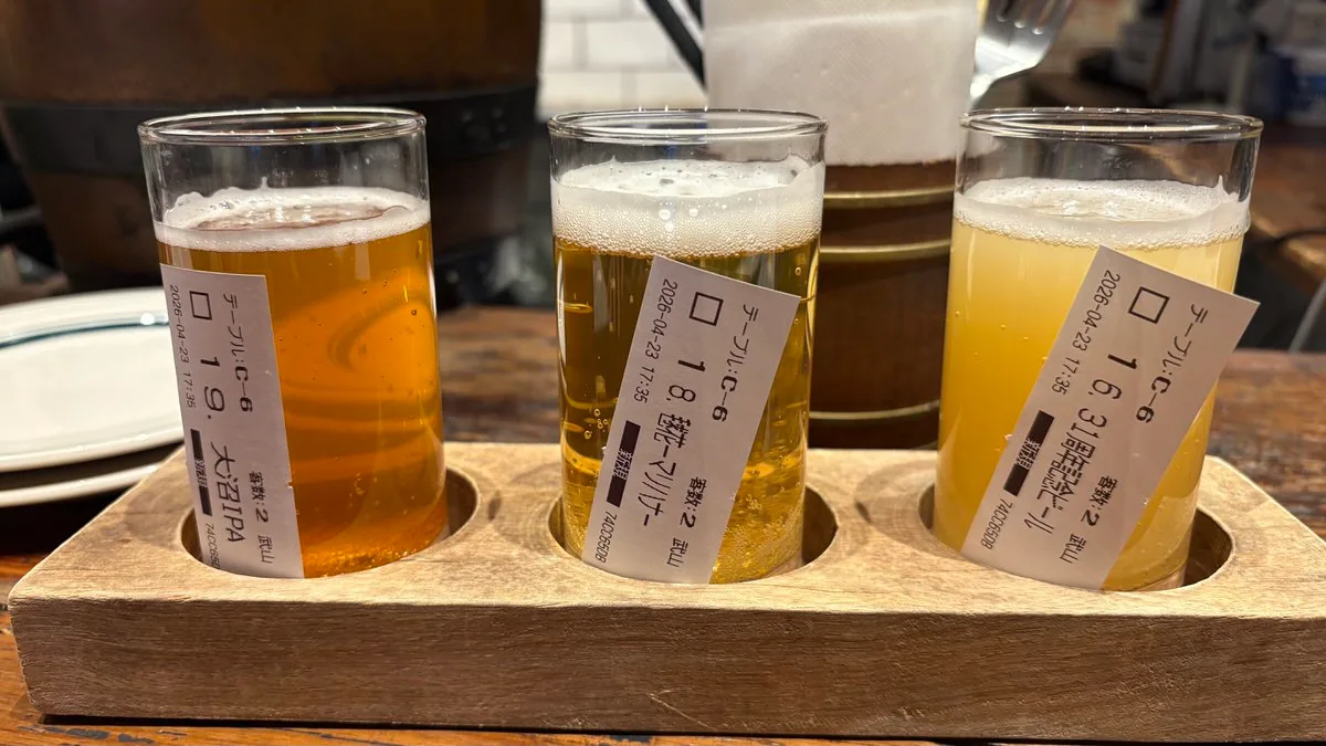 Three small glasses of beer in a wooden flight tray, each with a paper order slip taped to it