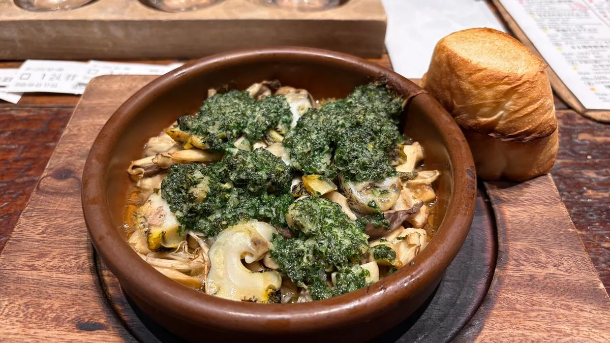 An earthenware dish of whelks and mushrooms under a green garlic and parsley topping, with a thick slice of toast beside it