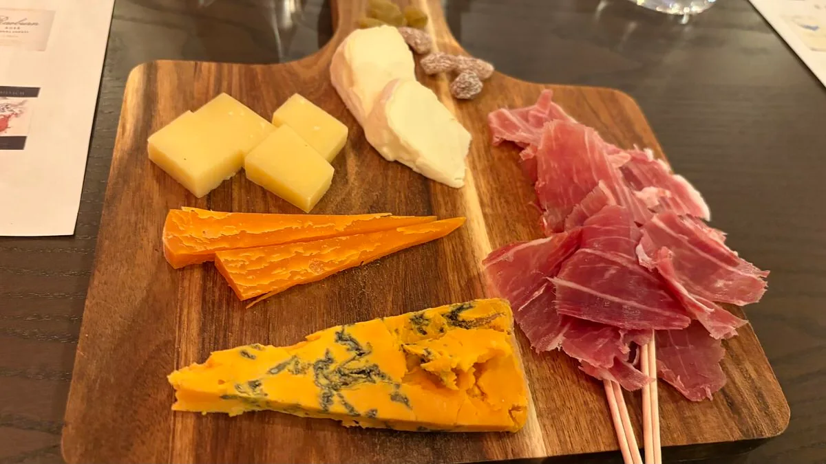A wooden board with cubes of pale semi-hard cheese, a white-rind soft cheese, slices of orange hard cheese, a wedge of orange blue-veined cheese, raisins and a pile of dry-cured ham on picks