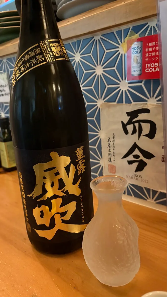 A black sake bottle with gold calligraphy beside a frosted glass carafe on a wooden counter
