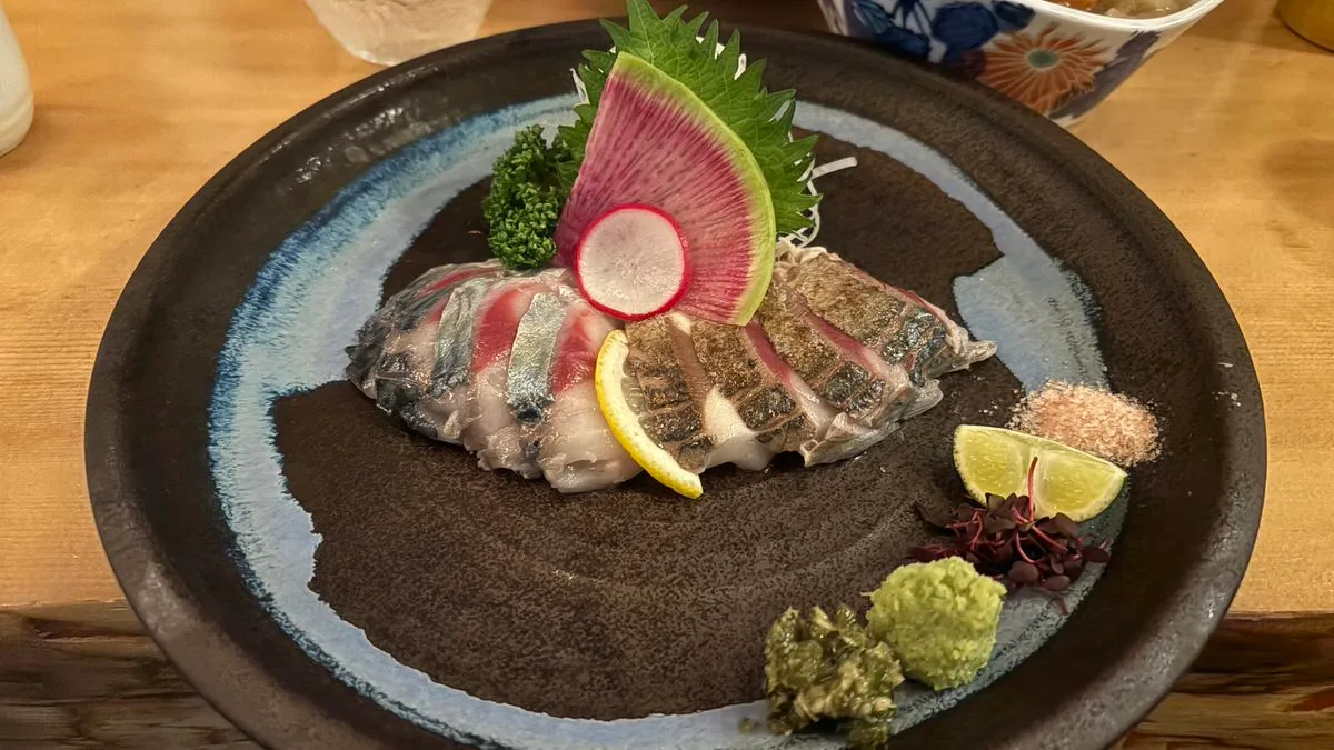 Slices of raw and seared mackerel arranged on a dark blue-rimmed plate with shiso, radish, wasabi, salt and citrus wedges