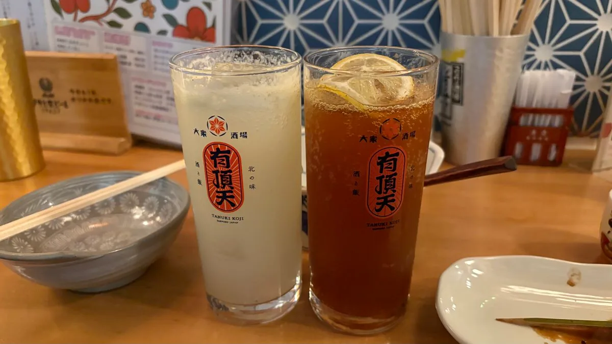 Two tall glasses printed with the shop's red logo, one pale and cloudy, one amber with a slice of lemon floating in it