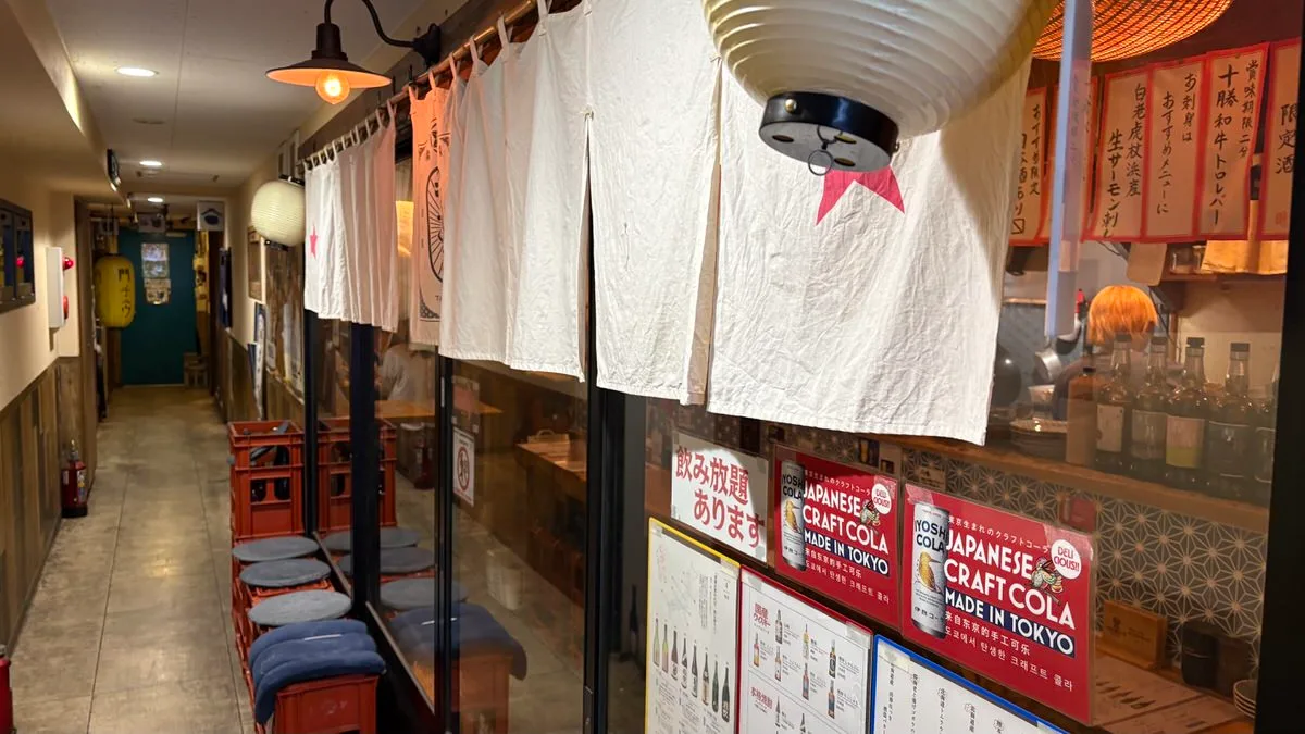 A narrow corridor with white noren curtains hanging along it, paper lanterns overhead and handwritten red signs on the wall