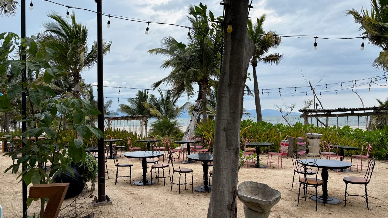 Round black tables and pink and black metal chairs on sand under strings of light bulbs, with palms and the sea behind