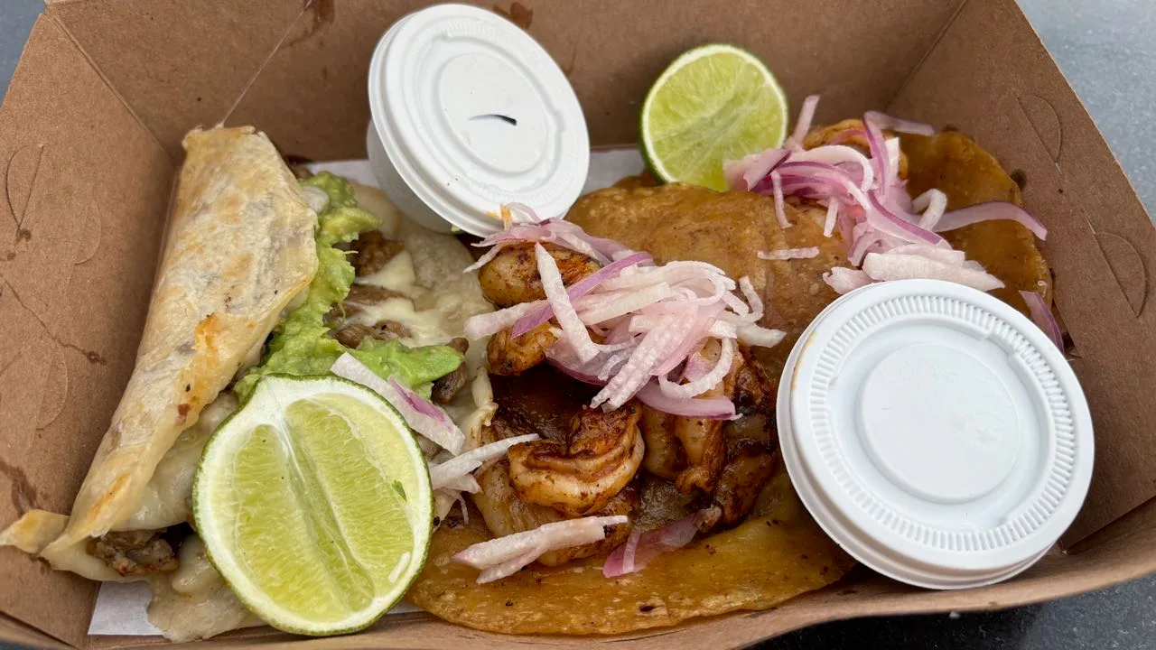 Two shrimp tacos on corn tortillas under slivers of red onion, beside a folded quesadilla with avocado and melted cheese, with pieces of lime and two lidded salsa cups in a cardboard box