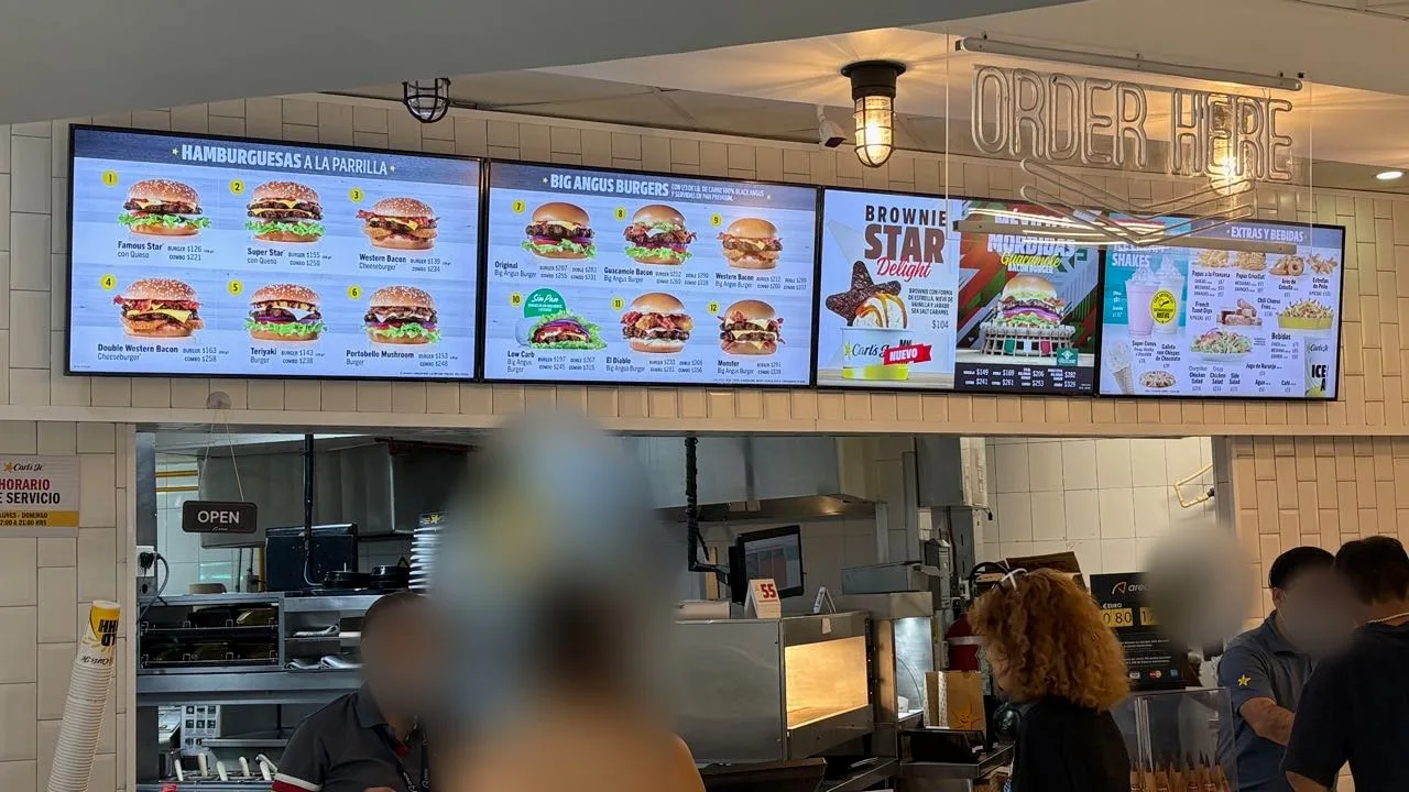 Digital menu boards above a Carl's Jr counter headed Hamburguesas a la Parrilla and Big Angus Burgers, with burger and combo prices, under a neon Order Here sign