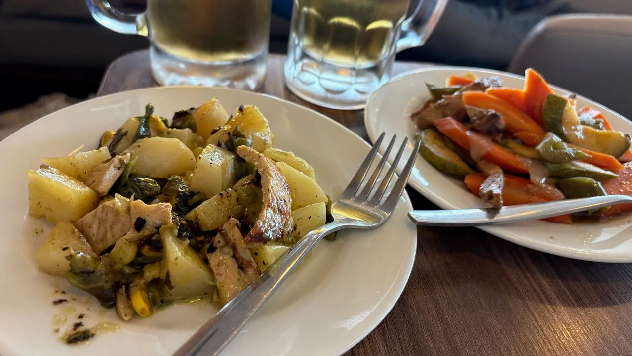 A white plate of chicken with potato chunks and green vegetables, a second plate of carrot, courgette and green pepper strips under a pale sauce, two glass mugs of beer behind them