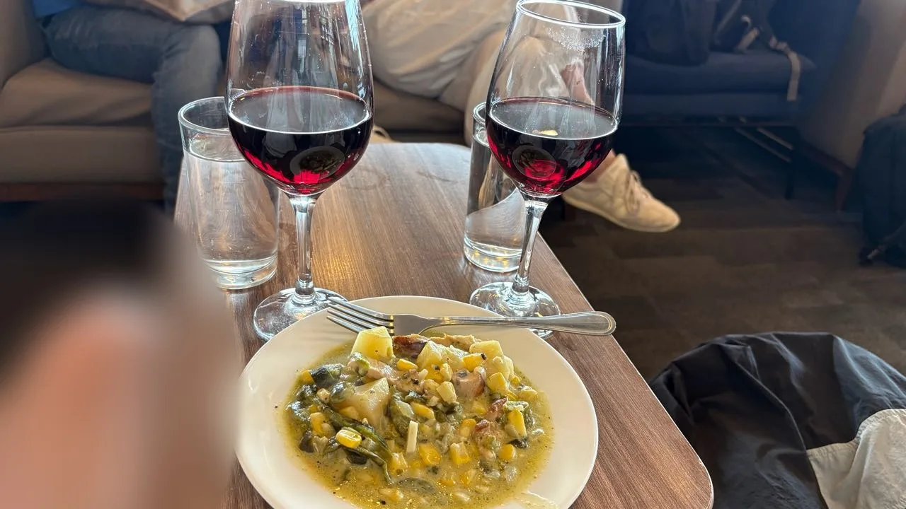 A plate of chicken with corn, potato and green vegetables in a light sauce on a wooden table, with two glasses of red wine and a glass of water behind it