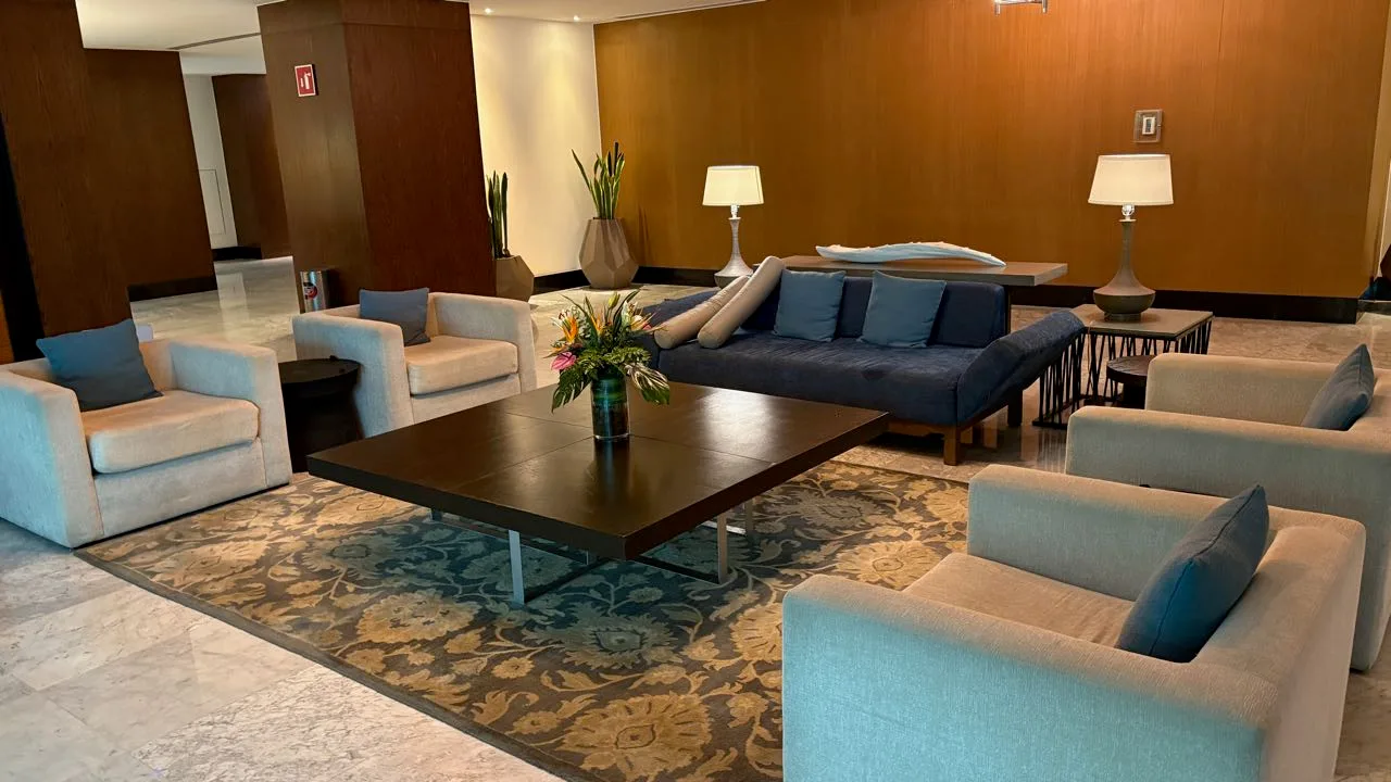 An empty lobby seating area with pale armchairs, a navy sofa, a low wooden table with a vase of flowers and a patterned rug