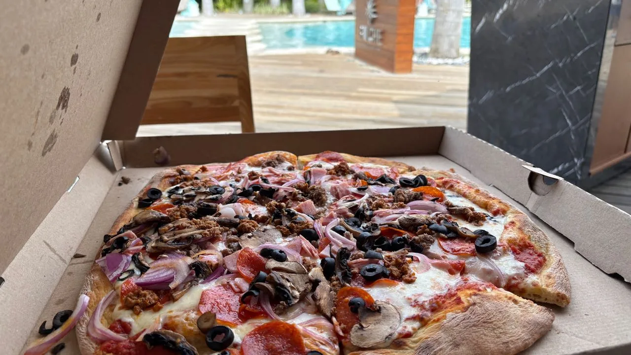 An open pizza box with a large pizza topped with pepperoni, black olives, mushrooms, red onion and ground meat, a pool and a wooden sign behind it