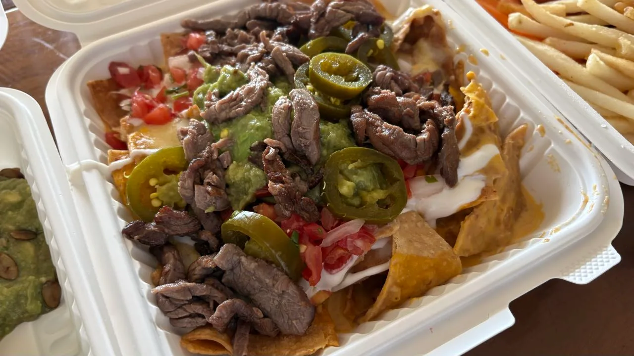 Tortilla chips in a takeout box under strips of grilled beef, jalapeño slices, guacamole, pico de gallo and sour cream, with fries and a seed-topped guacamole at the edges