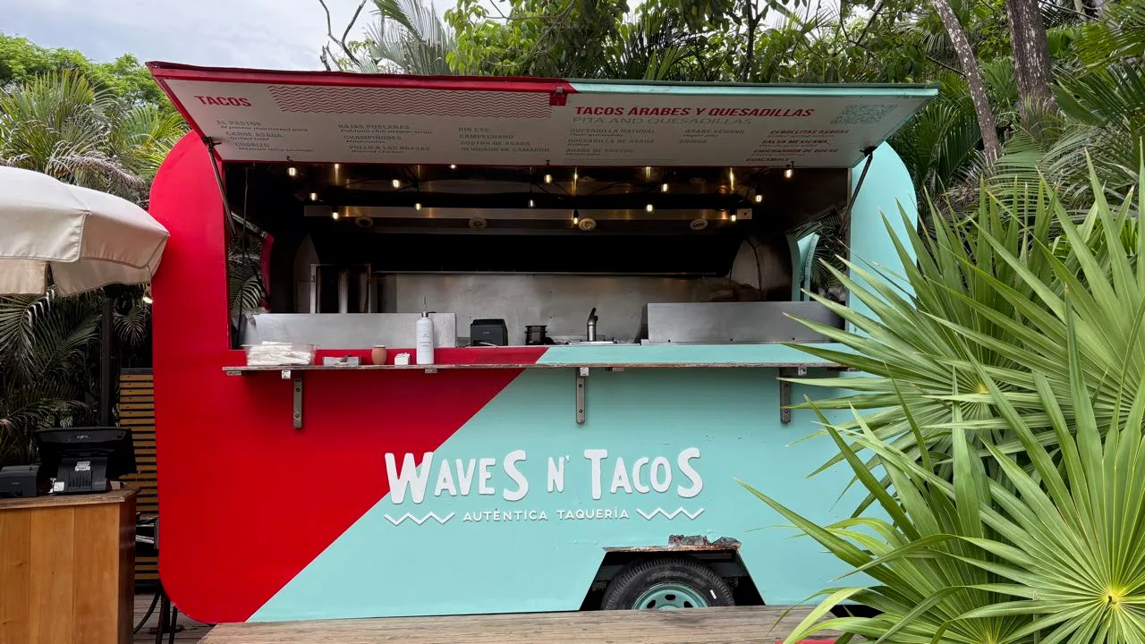 A red and turquoise food truck painted Waves N' Tacos, Auténtica Taquería, its raised hatch showing a menu board of dish names