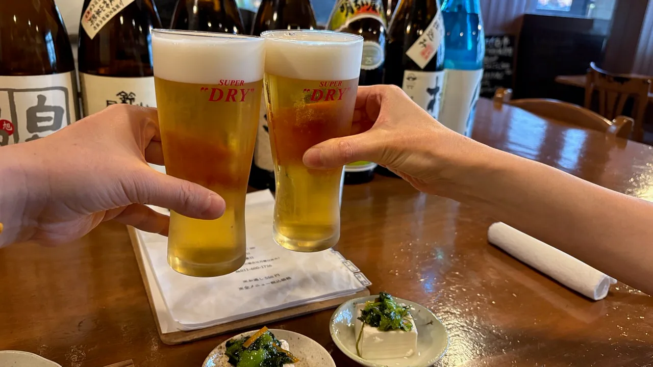 Two glasses of Asahi Super Dry draft beer being clinked over a wooden counter, with small dishes of cold tofu topped with green pickle