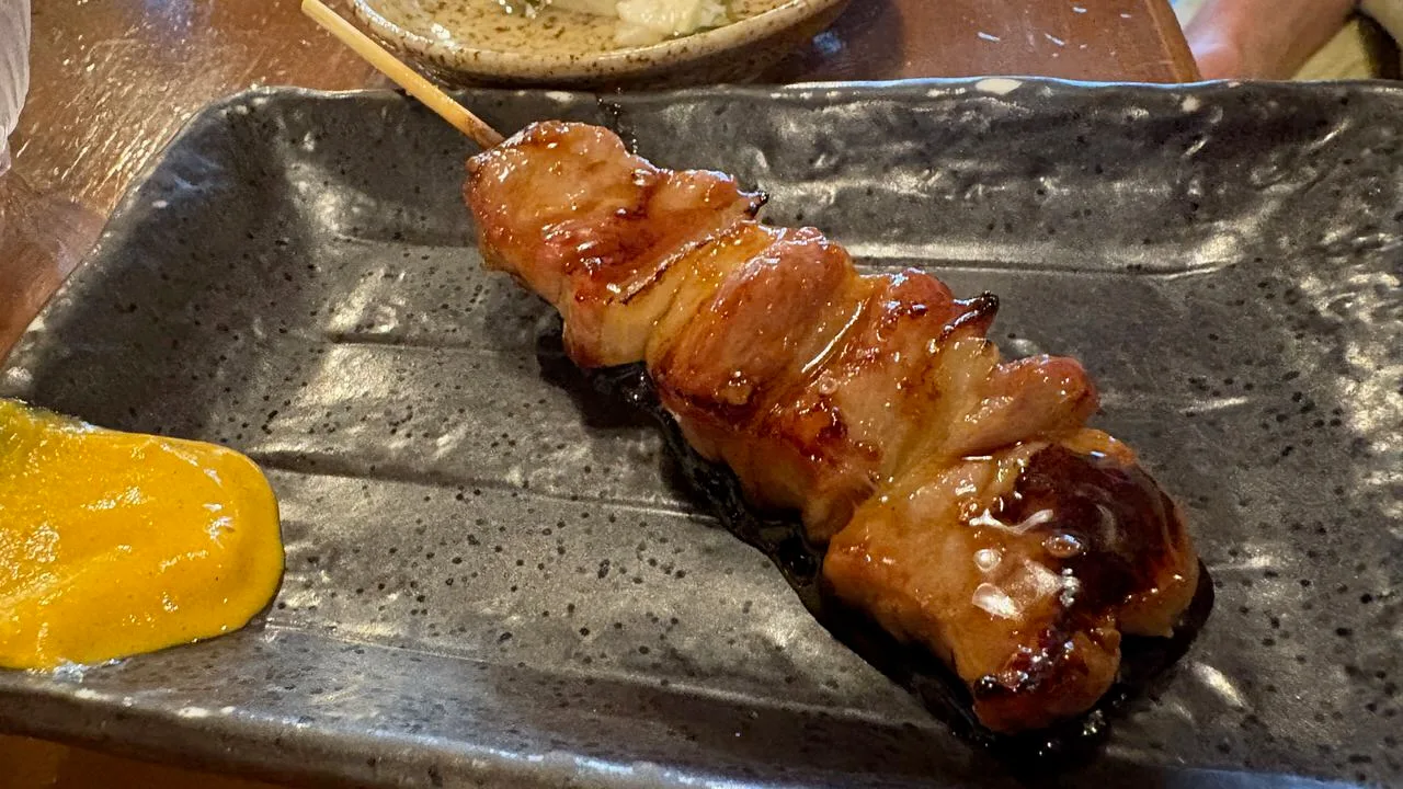 A single skewer of glazed grilled pork on a dark speckled plate with a smear of yellow mustard beside it