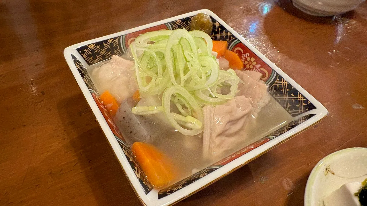 A square patterned bowl of offal (motsu) simmered in clear broth with daikon, konnyaku and carrot, topped with sliced spring onion