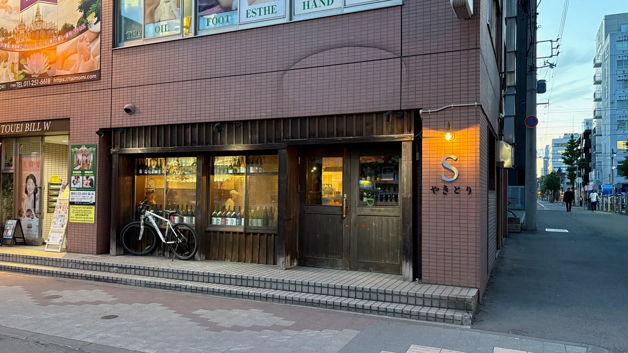 A corner shopfront at dusk with a wooden facade, bottles lined up in the window and a metal letter S on the wall